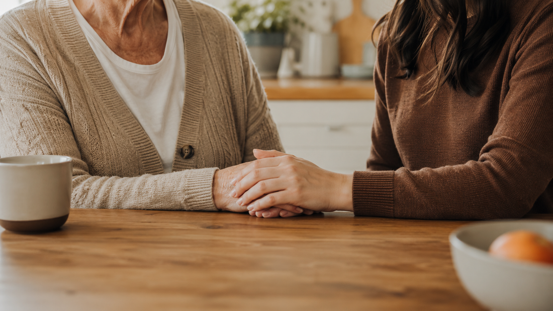 An adult daughter sits close beside her elderly mother at a kitchen table, gently holding her hand, representing the quiet, ongoing caregiving that defines life with Alzheimer's disease.