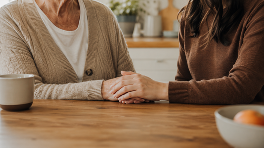 An adult daughter sits close beside her elderly mother at a kitchen table, gently holding her hand, representing the quiet, ongoing caregiving that defines life with Alzheimer's disease.